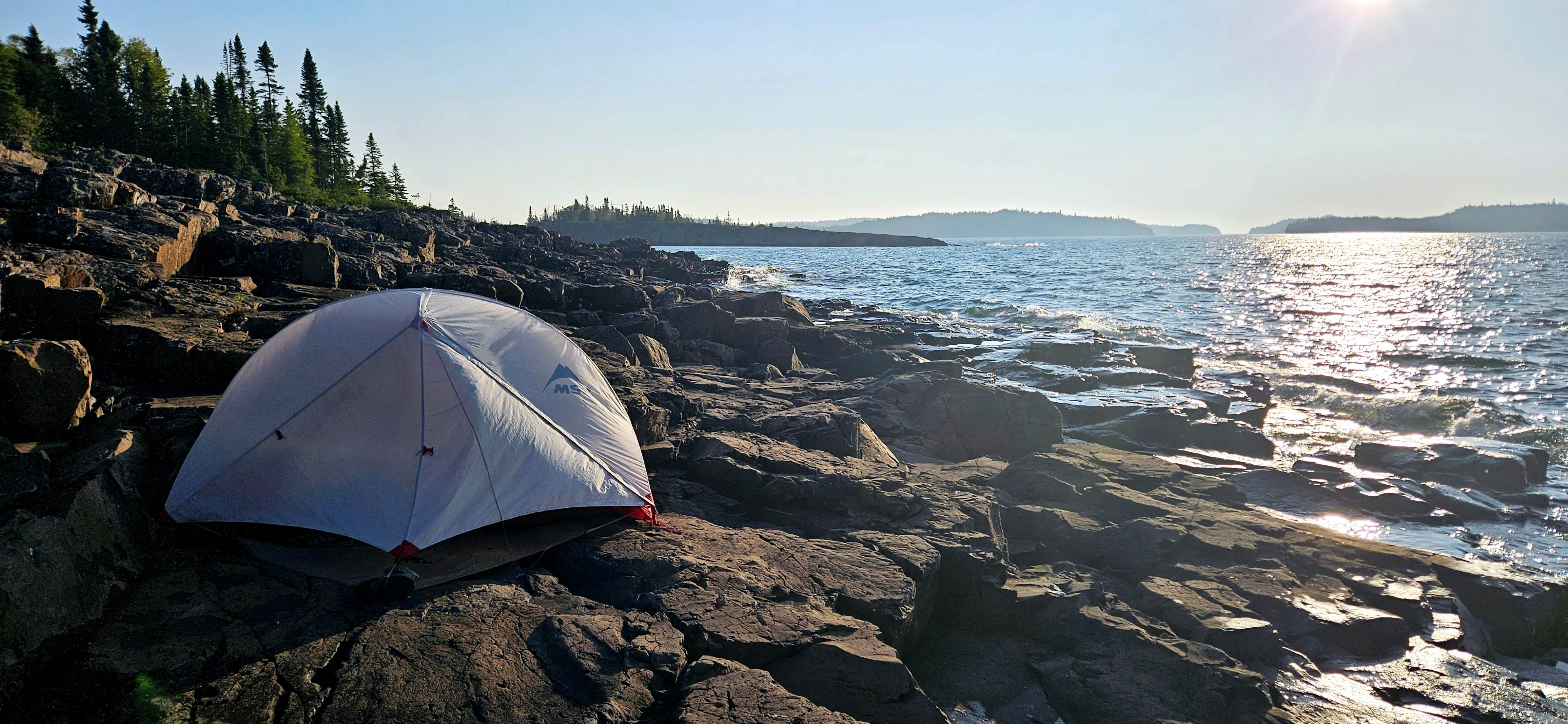 A tent sits on a rocky lakeside. The water stretches out across the right side of the photo, fading in to a blue sky.